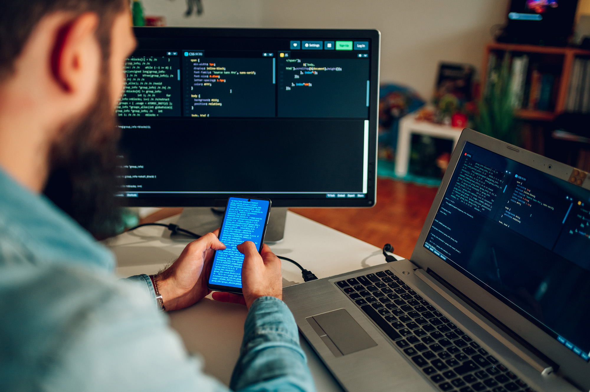 Rear view of an IT expert checking on data on his phone while sitting at his home office surrounded by computer screens with data and codes on them. Focus on the programmer's hands holding a phone.